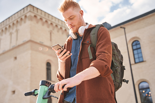 mann med skjegg, sekk og sparkesykkel ser på mobiltelefonen