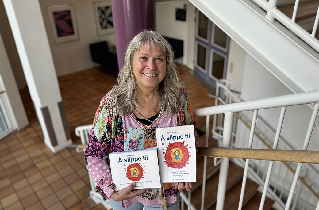 Bildet viser forsker May Østby stående i en trapp på campus med de to versjonene av boken på armen.