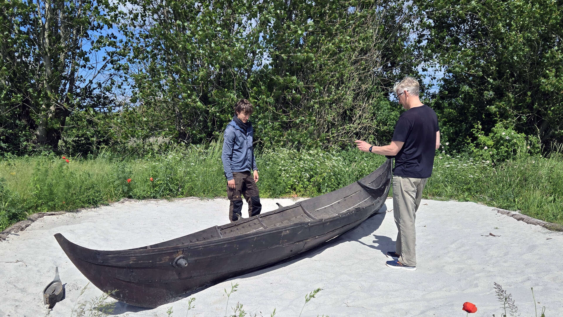 Two men examining a traditional, dark wooden small boat resting on a patch of white sand. The area is surrounded by green grass, red poppies, and trees under a clear blue sky.