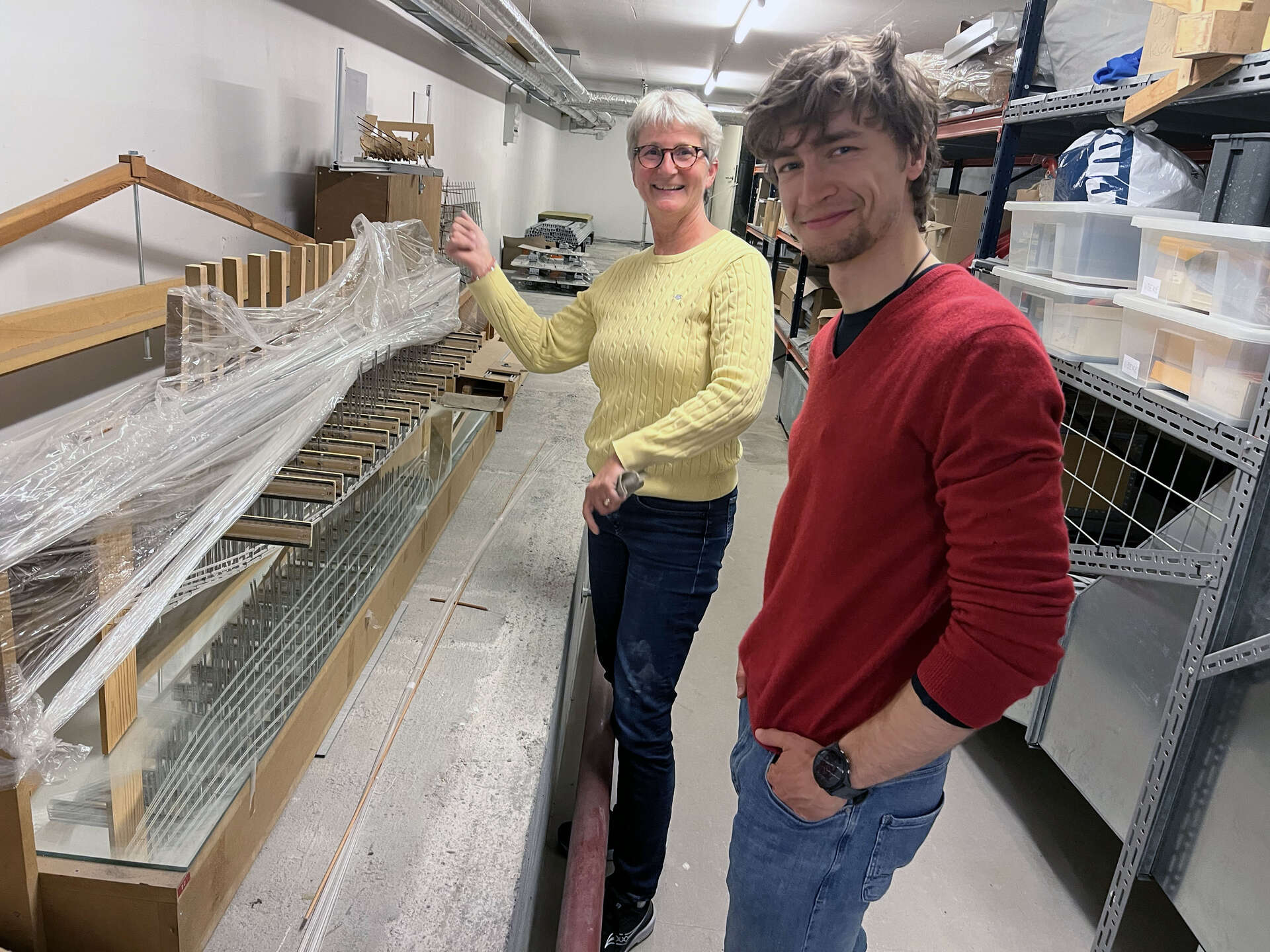 A woman in a yellow sweater and a young man in a red sweater standing in a basement or storage room, smiling at the camera. Next to them is a long wooden structure covered in plastic wrap, and shelves filled with boxes are visible in the background.