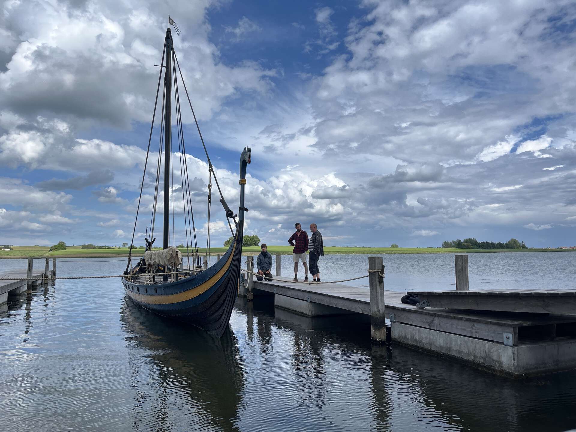 A traditional Viking longship moored alongside a wooden floating dock on a calm body of water. Three people are standing on the pier next to the boat, with a green landscape and a dramatic, cloudy blue sky in the background.