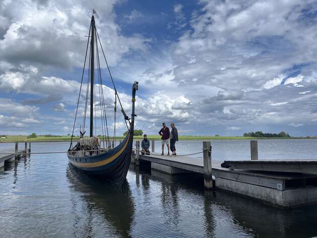 A traditional Viking longship moored alongside a wooden floating dock on a calm body of water. Three people are standing on the pier next to the boat, with a green landscape and a dramatic, cloudy blue sky in the background.