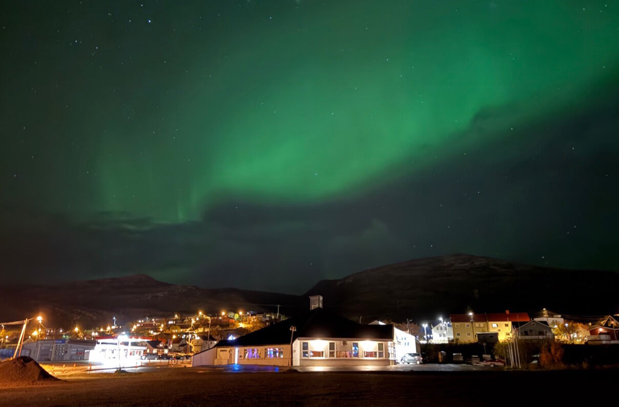 Bildet er tatt utendørs og viser en hvit, lav bygning med nordlys på himmelsen over.