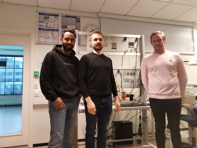De tre prosjektdeltakerne står i et laboratorium på høgskolen med vindturbinsimuleringsutstyr i bakgrunnen.