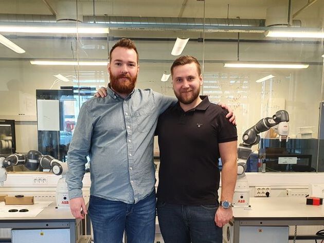 De to prosjektdeltakerne Johannes og Dennis står foran to montere med robotarmer i.
