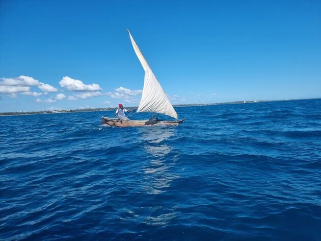 The photo shows a man in his boat at the sea. The sky is blue with only a few clouds. There are som small waves in the sea.