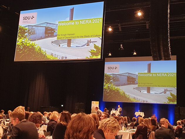 Bildet viser i nedkant en del av deltakerne på konferansen. Midt og øverste del av bildet viser to store digitale skjermer med bilde og teksten "Welcome to NERA 2021"  blant annet.