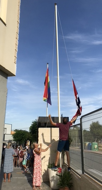 Et norsk flagg fires på en flaggstang. I bakgrunnen ses et Pride-flagg.