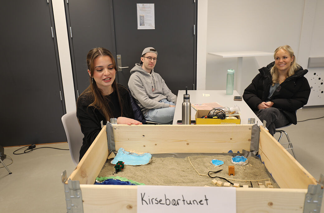 Bilde viser tre studenter som diskuterer og prater rundt en pallekasse i et klasserom. I pallekassen er et etablert et mikrosamfunn som studentene har ansvaret for å utvikle. 
