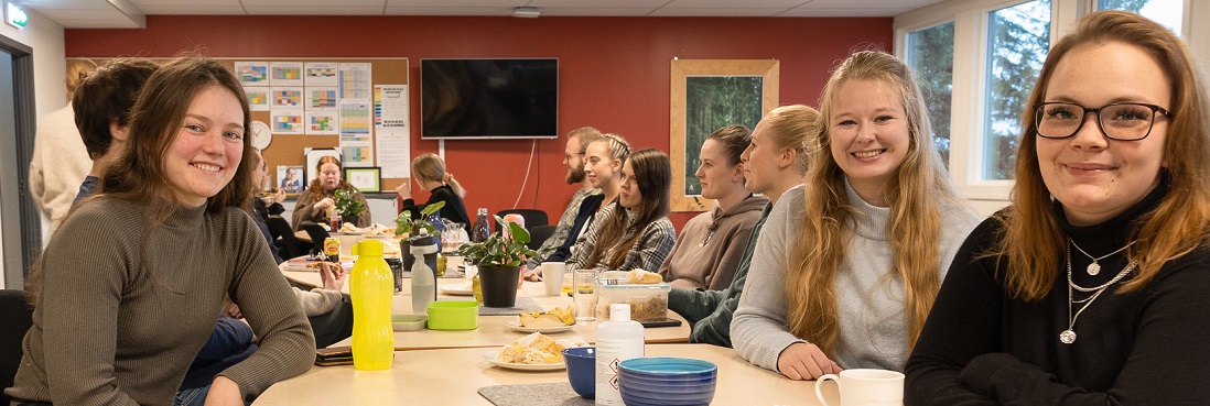 Flere personer sitter rundet et bord. De tre fremste ser inn i kameraet.