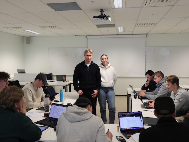 Bildet viser en gruppe studenter samlet i et klasserom sammen med to mentorer som står sentrert i bildet. 