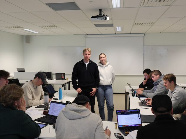 Bildet viser en gruppe studenter samlet i et klasserom sammen med to mentorer som står sentrert i bildet. 