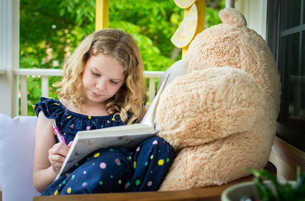 Et barn som skriver i en bok med en bamse vedsiden av seg