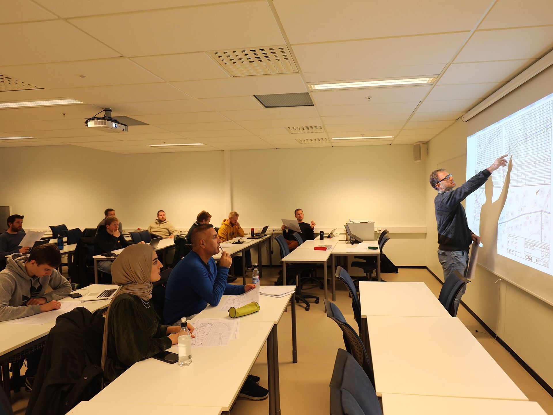 Foreleser foran tavle i et undervisningsrom med studenter. Foto.