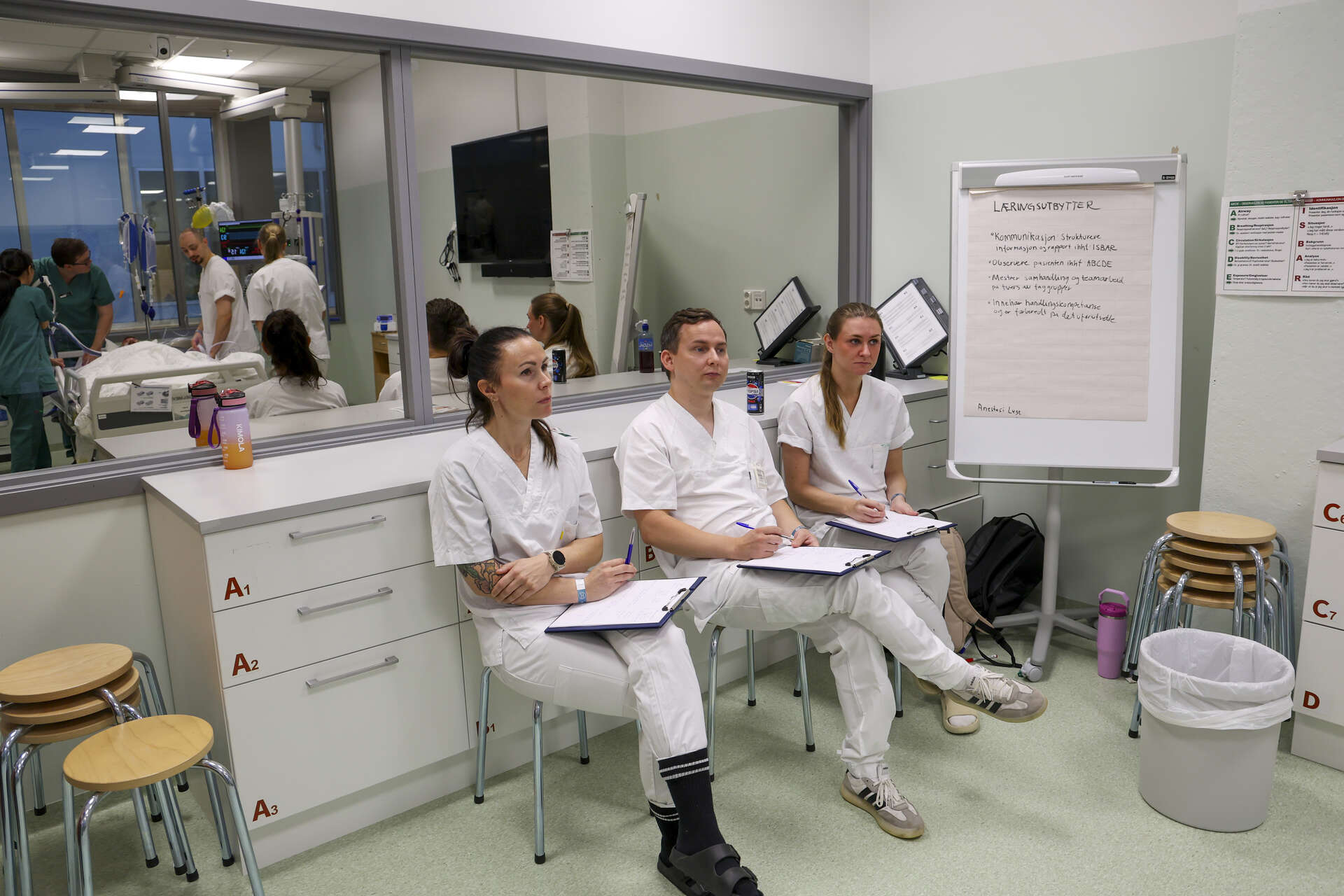 Tre sykepleierstudenter sitter og observerer simuleringen. I refleksjonen i vinduet bak dem ser vi andre studenter i gang med simulering.